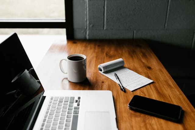 A laptop on a wooden desk with a notebook and a cup of coffee.